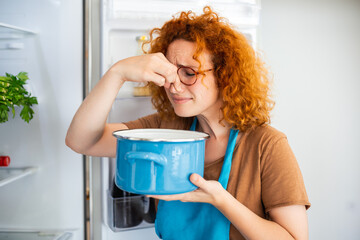 Bad Food In Fridge, young woman in holding her nose because of bad smell from food in refrigerator at home - obrazy, fototapety, plakaty