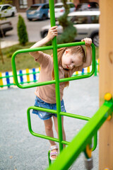 a child girl on a children's multi-colored playground in the courtyard of a multi-storey house in the summer has fun playing and climbing high up the stairs, summer holidays