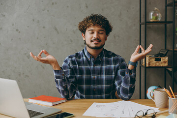 Successful employee business Indian man he wears casual blue checkered shirt spread hand in yoga om aum gesture relax meditate try to calm down sit work at office desk with laptop pc computer indoors