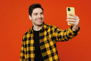 Young man he wear yellow checkered shirt black t-shirt doing selfie shot on mobile cell phone post photo on social network isolated on plain red orange background studio portrait. Lifestyle concept.