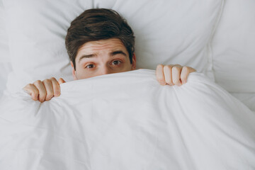 Close up young frightened scared sad man wear casual clothes white t-shirt pajama lying in bed look...
