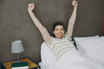 Young happy fun energetic man wearing casual clothes t-shirt pajama lying in bed do hands stretching rest relax spend time in bedroom home in own room hotel wake up dream be in reverie good mood day.