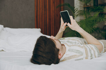 Side view young man wear casual clothes t-shirt pajama lying in bed hold use blank screen mobile cell phone rest relax spend time in bedroom home in own room hotel wake up dream be in good mood day.