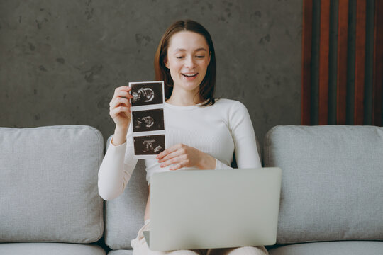 Young pregnant IT woman in casual clothes use laptop pc computer show ultrasound scan sit on grey sofa couch stay at home hotel flat rest relax spend free spare time in living room Maternity concept