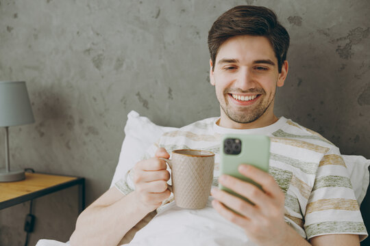 Young Fun Man Wear Casual Clothes T-shirt Pajama Lying In Bed Drink Coffee Tea Use Mobile Cell Phone Rest Relax Spend Time In Bedroom Home In Own Room Hotel Wake Up Dream Be In Reverie Good Mood Day.