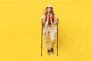 Full body smiling young woman carry bag with stuff mat hold trekking poles isolated on plain yellow background. Tourist leads active lifestyle walk on spare time. Hiking trek rest travel trip concept.
