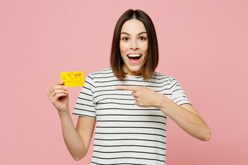 Young smiling happy fun woman wears casual clothes t-shirt hold in hand point finger on mock up of credit bank card isolated on plain pastel light pink background studio portrait. Lifestyle concept.