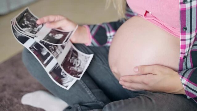 Woman sitting on the floor and stroking her naked pregnant belly, mom holding ultrasound scan pictures of her baby child gender reveal in hands, ultrasound medical examination of prenatal period