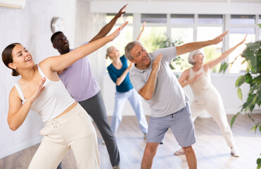 Fototapeta premium In fitness studio, youthful female coach is leading class for seniors. Instructor is teaching older students fundamental steps of hip hop dance in dance lesson