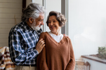 A couple of mature spouses cuddle nicely near a large window. An older man hugs his attractive...