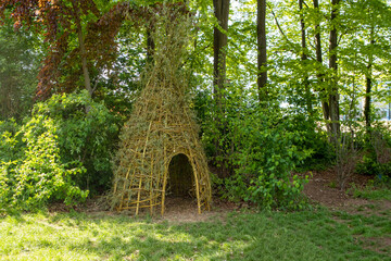 Brown willow teepee stands in front of green trees in a meadow