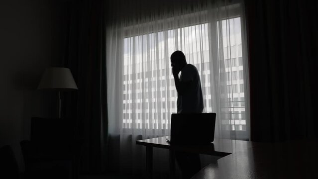 Silhouette Of Male Pacing Up And Down Beside Large Backlit Window In Hotel Room Talking Of Mobile And Gesturing At Laptop On Hotel Desk. Locked Off