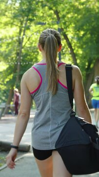 Vertical Screen: Woman with ponytail wearing sportswear carrying bag in public park, blurred people on background jogging, walking with dog and playing ping pong. Concept of healthy lifestyle