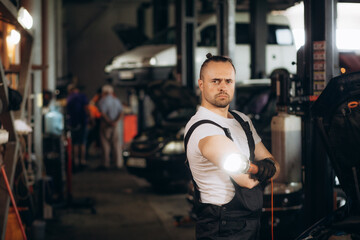 Handsome Car Mechanic is Posing in a Car Service. He Wears a Jeans Shirt and Safety Glasses. His Arms are Crossed. Specialist Looks at a Camera and Smiles