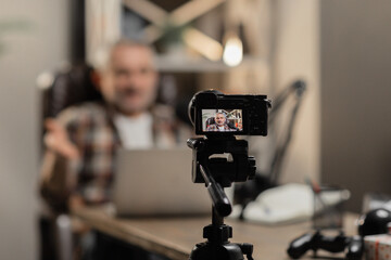 A small camera on a tripod takes pictures of a bearded man sitting at a table. A male blogger is recording a video using a camera standing on a table.
