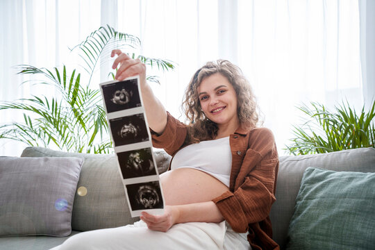 Smiling pregnant woman showing ultrasound scans sitting on sofa at home