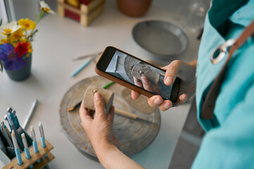 Craftswoman photographing clay cup through smartphone at workbench