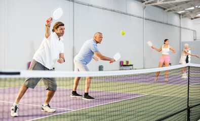 Portrait of sporty adult man playing doubles pickleball with experienced aged partner on indoor court, ready to hit ball. Sport and active lifestyle concept
