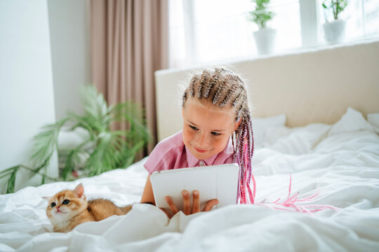 Girl holding tablet PC lying by ginger kitten on bed