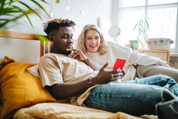 Smiling young woman looking at boyfriend's smart phone relaxing on bed