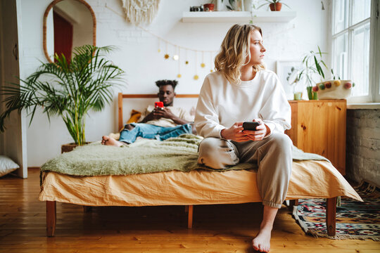 Thoughtful sad woman on bed with boyfriend using phone in background