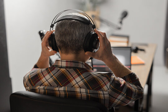 A man in a plaid shirt sits at a table in front of a laptop and a microphone. A man with gray hair puts on headphones and prepares to record a new podcast. - Powered by Adobe