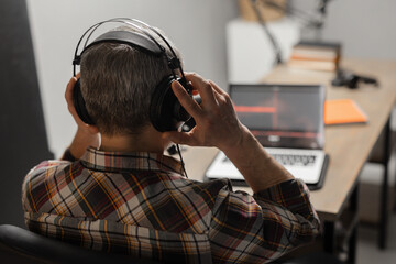 A gray-haired man with headphones on his head sits at a table in front of a laptop. A male blogger adjusts the headphones on his head while recording a podcast.