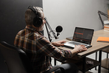 A man in headphones records a podcast using a studio microphone and a laptop. A gray-haired man in a plaid shirt sits at a table and looks at a computer screen.