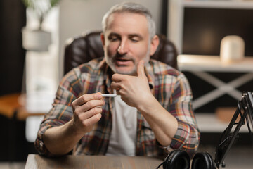 A bearded man in a plaid shirt sits at a table and looks thoughtfully at a cigarette in his hand.