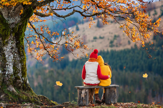 Hello Autumn. Children Are Sitting On A Wooden Bench Under A Old Tree With Autumn Leaves. Family Walk In Autumn.