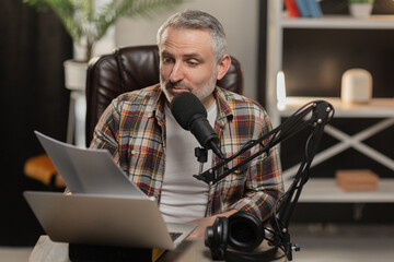 A bearded man sits at a table in front of a laptop and prepares to record a video. A male blogger is sitting at his desk carefully rereading the script for his new video.
