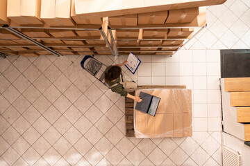 Top view of worker checking merchandise inventory on laptop computer, preparing customers orders for delivery in storehouse. Storage room employee wearing industrial overall in warehouse