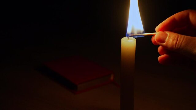 Close-up of a man's hand lighting a wax candle with a match against a black background. The bright flame of a candle illuminates an ancient book lying on the table. Symbol of memory, ritual, ceremony