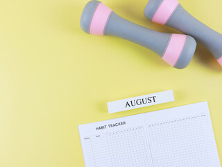 flat lay of habit tracker book, wooden calendar August, gray pink dumbbells,  on yellow  background with copy space. Self development and active lifestyle concept.