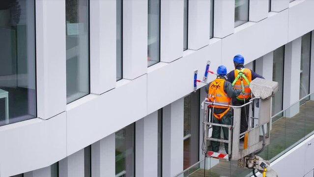 Two workers wearing safety harness wash office building facade at