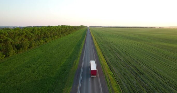 Semi trucks drive along the highway, aerial shot. Freeway truck traffic at sunset in summer. Green field. Drone view. Logistics industry. Cargo transportation
