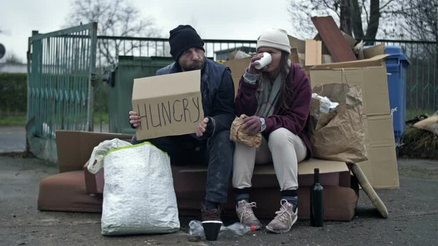 Two dirty and poorly dressed homeless people, a man and a woman, sit by a pile of rubbish with a handwritten HUNGRY poster. Woman drinking something from a disposable glass.