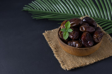 Dark dried dates lie in a wooden bowl on a canvas napkin. Green basil leaf on top. Palm leaf on a dark table. Dark background. Space for text