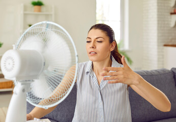 Woman suffering from summer heat and using electric fan to refresh in current of cool air. Young...
