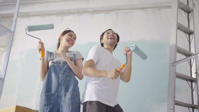 Young Asian couple Dancing together while repairing and painting the wall with blue paint using a roller during renovation in their new apartment.