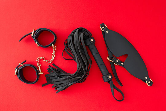 Concept of BDSM toys for erotic indulgence. Top view arrangement of handcuffs, blindfold, leather whip on red background
