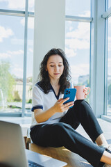 Smiling curly-haired woman sitting in a loft cafe, working at a computer, reading messages on a smartphone. Remote work