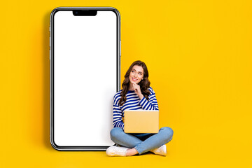 Full body photo of minded positive lady sit floor use netbook hand touch chin look empty space huge telephone isolated on yellow color background
