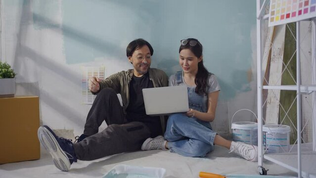 Young Asian couple repairing and painting the wall Sit and look at home decorating ideas through a laptop.
