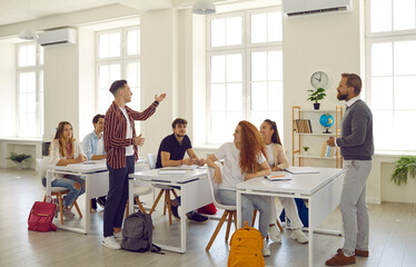 Group of high school students having a very interesting lesson in a modern classroom. Male student speaks up in class. Young man stands up and answers a question as his classmates and teacher listen