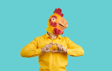 Funny man with comic romantic mood wearing chicken mask on his head shows heart shape showing love intentions. Eccentric man in orange shirt with absurd rubber mask chicken on light blue background.