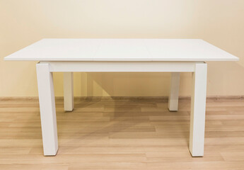 A white table with four legs stands in the interior of a room with a laminate floor and plain walls.