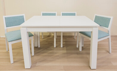 A white table with four legs stands in the interior of a room with a laminate floor and plain walls; there are four chairs with turquoise upholstery near the table.
