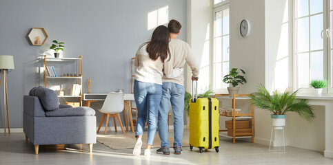 Happy couple in love back home from summer holiday trip. View from behind of young man and woman who have just returned from vacation travel with yellow suitcase standing in living room and hugging