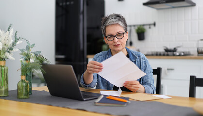 Senior woman at a table with a laptop in a modern interior holds reports by mail, documents in her...